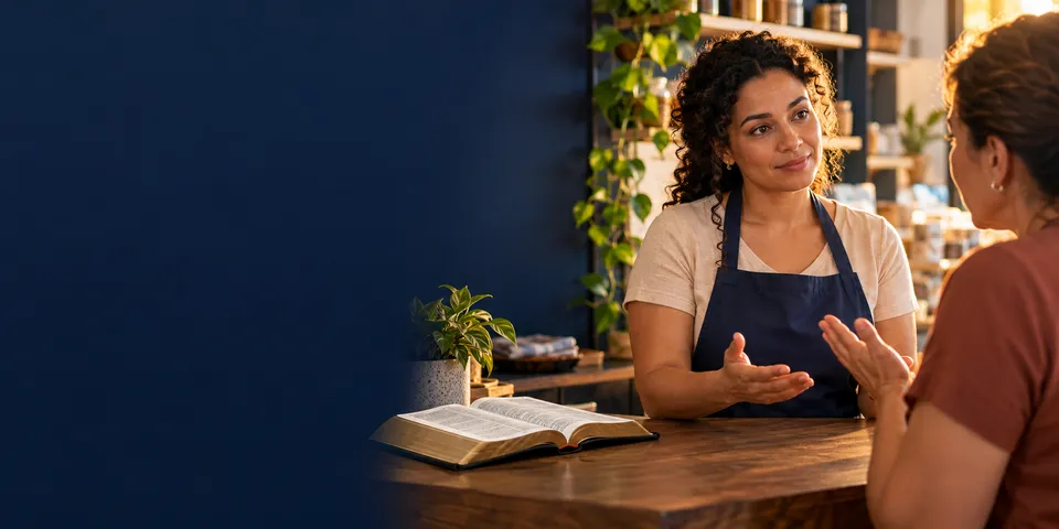 Mulher de avental conversa com calma com outra pessoa, gesticulando, à mesa com uma Bíblia aberta.