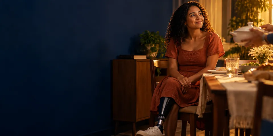 Mulher com uma prótese na perna, sentada à mesa de um jantar em família, sorri enquanto alguém lhe serve um prato.